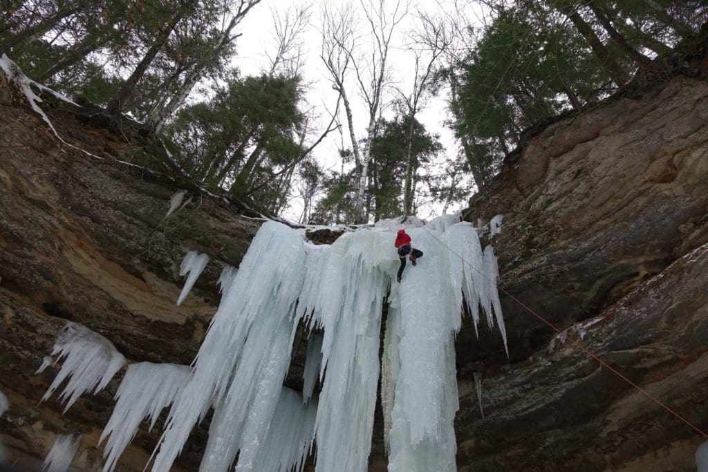 Winter in the Parks 20 Ice Climber near the top of a curtain and column of ice. The ice is forming out of and hanging off sandstone rocks. Trees are on top of the cliff.